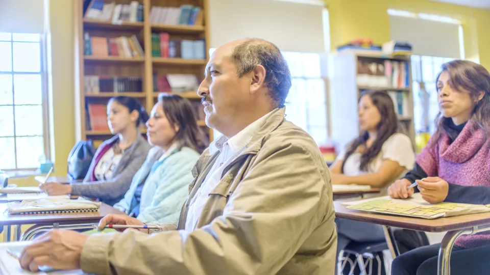 Kommunerna måste bli bättre på att följa upp kvaliteten på komvux på entreprenad Vuxna studenter sitter i ett klassrum och lyssnar uppmärksamt, med anteckningsblock och pennor framför sig, omgivna av bokhyllor och fönster som släpper in dagsljus.