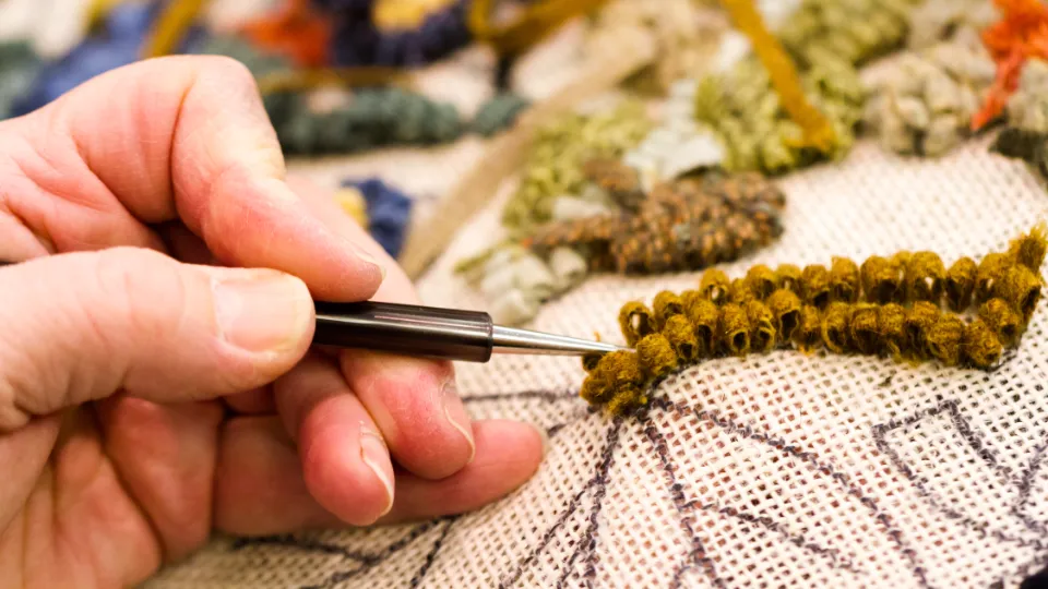 Lärare undervisar på äldreboende Närbild av en hand som använder en punch needle-teknik för att brodera mönster med brunt garn på ett tyg med konturteckningar.