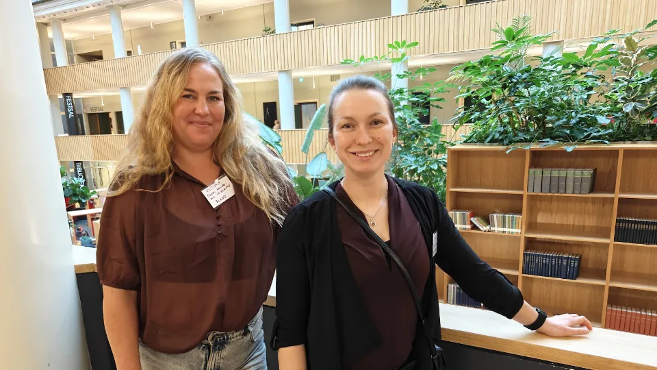 Victoria Nylund og Linda Heinola smilende foran en bogreol i et lyst og moderne kontormiljø med grønne planter i baggrunden.