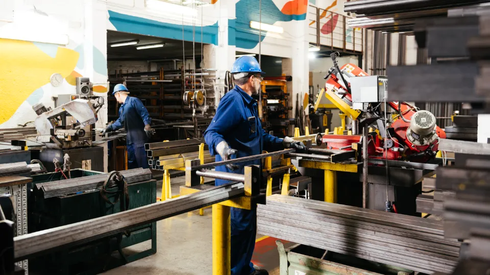 Industriarbeidere får betalt permisjon for etter- og videreutdanning To industriarbeidere med verneutstyr jobber med metallbearbeiding i en fabrikkhall omgitt av maskiner og stålrør.