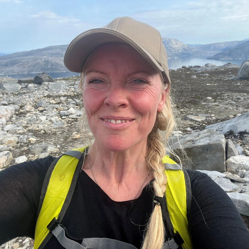 Lene Andersen iført kasket og rygsæk, smilende i fjeldlandskab med udsigt over vand og bjerge i Grønland.
