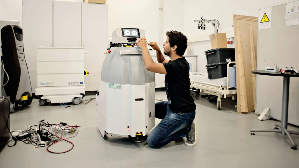 NLL i spidsen for nyt projekt om fremtidens kompetencer Mand arbejder på en service-robot i et laboratorium med teknisk udstyr og hospitalsseng i baggrunden.