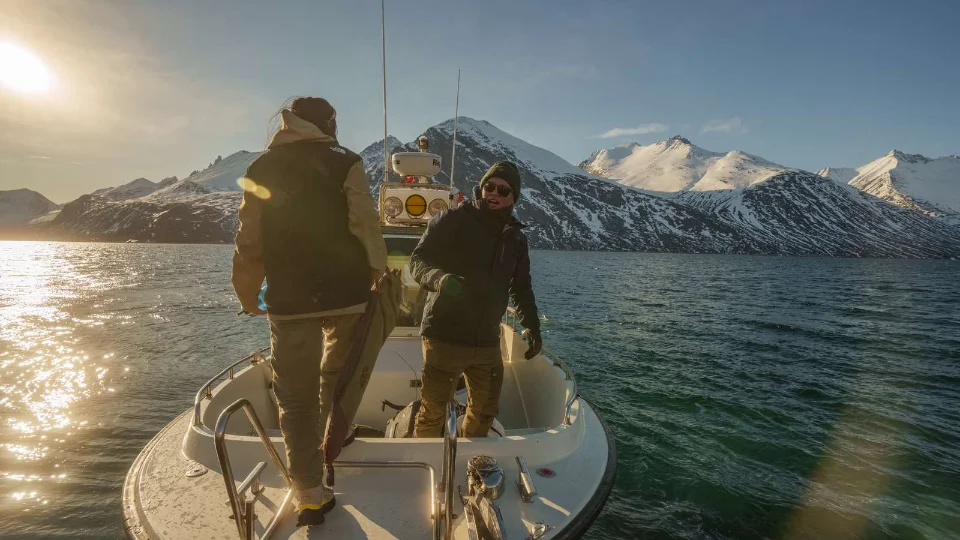 NEET To mænd på en båd i en fjord omgivet af snedækkede bjerge under en lav vintersol.