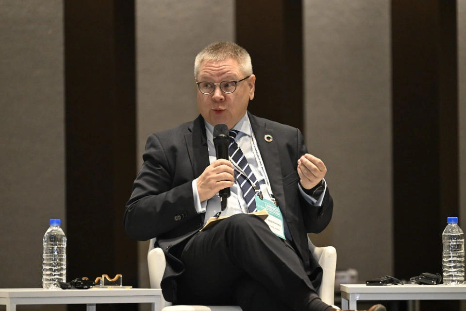 Rilli Lappalainen sitter i en stol under en konferens, håller en mikrofon och pratar engagerat, med vattenflaskor och anteckningar på borden bredvid.