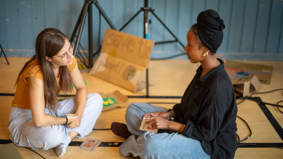 Samtale og fællesskab er kernen i folkeoplysning Två kvinnor sitter på golvet och samtalar, omgivna av skyltar och konstmaterial i en kreativ miljö.