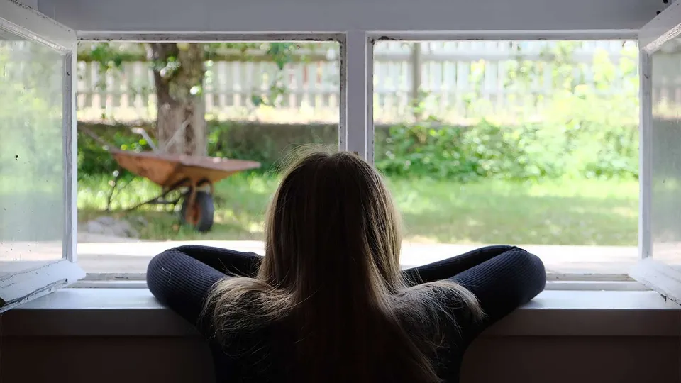 Education for Women in Norwegian Prisons Girl with long hair leaning on windowsill, looking out at a sunny garden with a wheelbarrow and tree in the background.