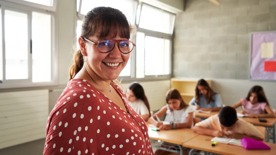 EPX-ekspertgruppen anbefaler nationalt kompetenceudviklingsprogram En smilende kvindelig lærer med briller og prikket bluse står i et klasseværelse, mens elever arbejder koncentreret ved deres borde i baggrunden.