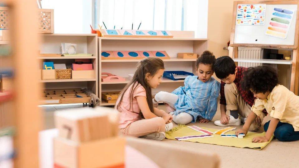 Förbättra kvaliteten på komvux och sfi Fyra barn i förskoleåldern sitter tillsammans på ett ljust golv och leker med färgglada pedagogiska verktyg. De fokuserar på en aktivitet med olikfärgade brickor som ligger utspridda på en liten matta. I bakgrunden syns ljusa trämöbler med prydligt sorterat material och en whiteboard, vilket skapar en inspirerande läromiljö.