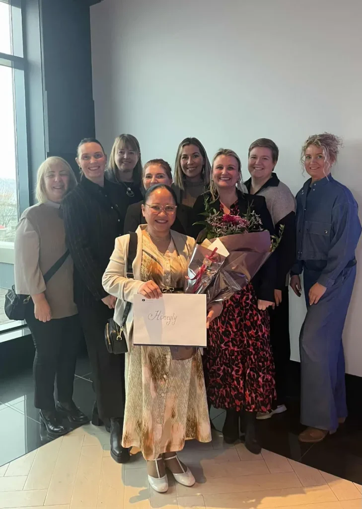 Gruppebillede af kvinder indendørs, hvor en kvinde i midten holder en buket blomster og et diplom foran en lys væg.