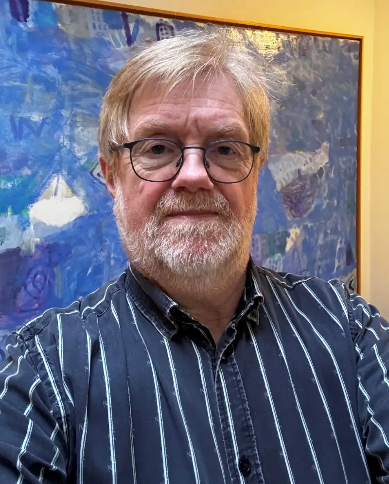 John Dalsgarð, der bærer briller og har lyst hår samt et velplejet gråt skæg. Han er iført en mørkeblå stribet skjorte og står foran et stort, farverigt abstrakt maleri med dominerende blå nuancer.
