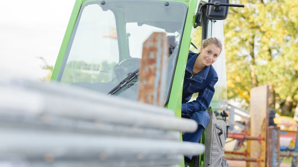 Satsing på fagskolene: tildeling av 900 nye studieplasser En smilende ung kvinne i blå arbeidsdress sitter i førerhuset på et lysegrønt anleggskjøretøy eller en gaffeltruck. Hun ser ut mot kameraet mens hun sitter ved rattet. I forgrunnen er det noen uskarpe metallrør, og i bakgrunnen kan man se trær med grønt løv.