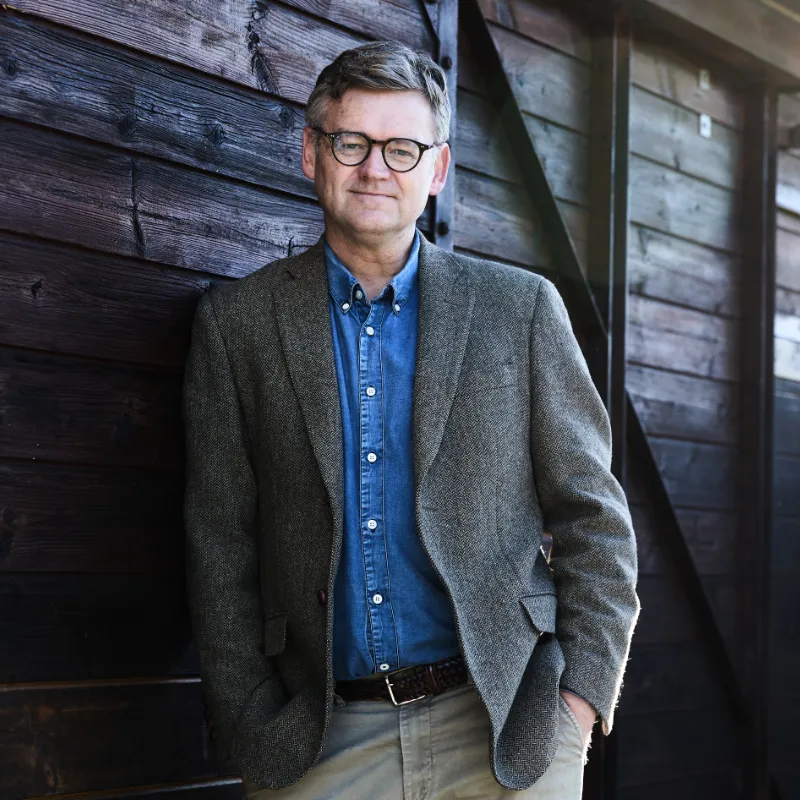 Rasmus Willig står afslappet op ad en mørk trævæg iført blazer, denimskjorte og briller.