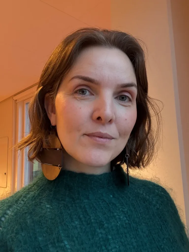 Synnøve av Rana ser direkte ind i kameraet med et mildt blik. Hun har mellemlangt brunt hår, bærer en mørkegrøn striktrøje og iøjnefaldende store, cirkelformede øreringe af træ. Baggrunden er enkel og oplyst af et varmt, blødt lys fra siden.