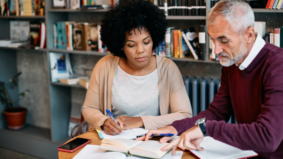 Universiteter skal varetage SPS-system for voksne studerende To personer studerer sammen ved et bord i et bibliotek, omgivet af bøger og noter.