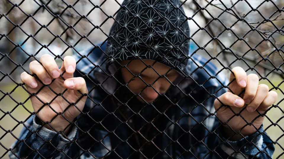 Fengsling av barn som siste utvei Ung mann med hette bøyer hodet ned og holder fast i et svart nettinggjerde.