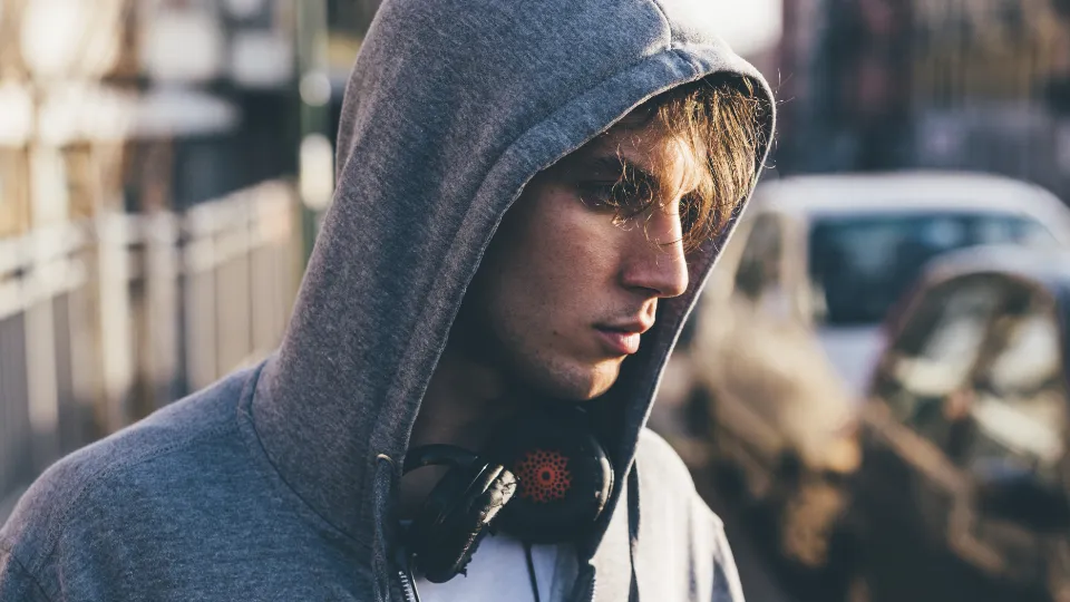Folkhögskolan hjälper unga i utanförskap mot studier och arbete Ung man i grå huvtröja med hörlurar runt halsen ser fundersam ut på en gata.