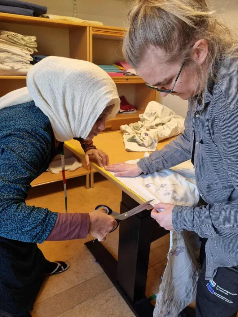 Två kvinnor står nedböjda över ett bord, den ena håller i tyget och den andra klipper.