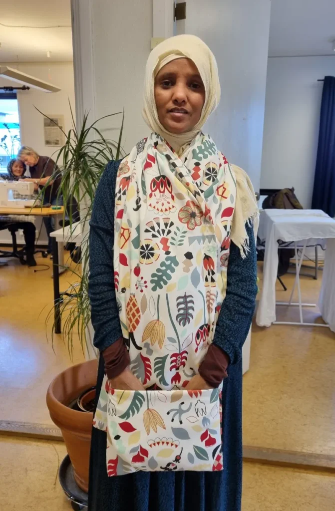 Yngre kvinna med ljus schal över håret, med blommigt tygstycke runt halsen som även täcker större delen av framsidan av hennes kropp.