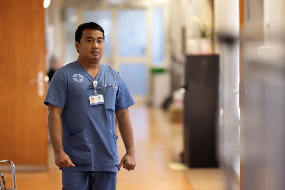 Mandlig sygeplejerske står på hospitalsgang iført blå uniform og navneskilt.