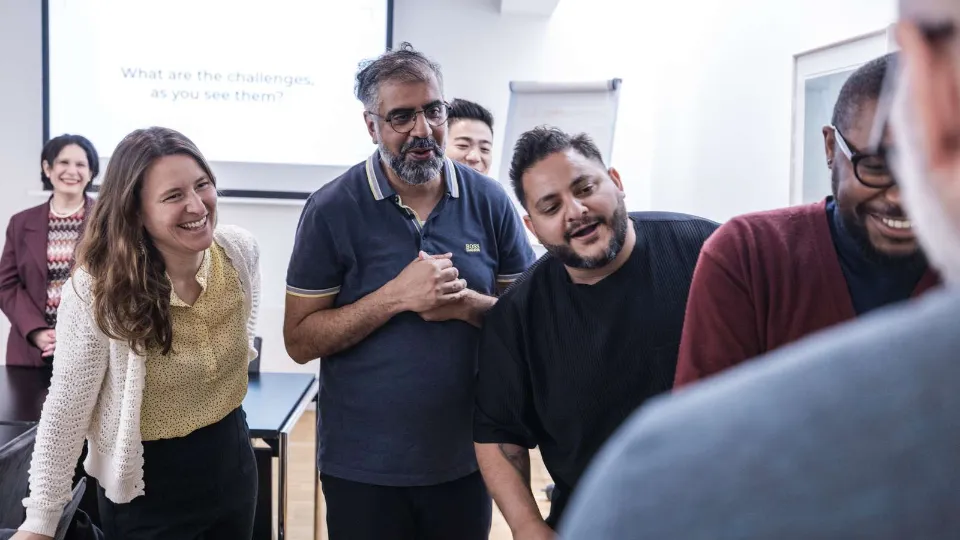 Læringscirkler er en gammel og moderne vej til udvikling Smilende kolleger i et mødelokale under workshop, hvor en gruppe diskuterer foran en projektorskærm.