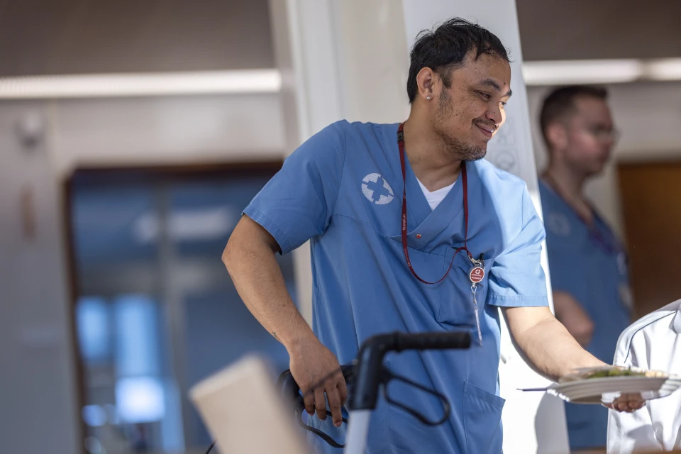 Mannlig helsearbeider i blå uniform serverer mad til patient på plejehjem.