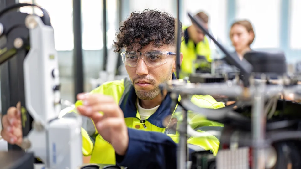 Regeringen förbättrar förutsättningar för yrkesutbildningar i Sverige Person i skyddsglasögon och varseljacka arbetar vid maskin i verkstadsmiljö.