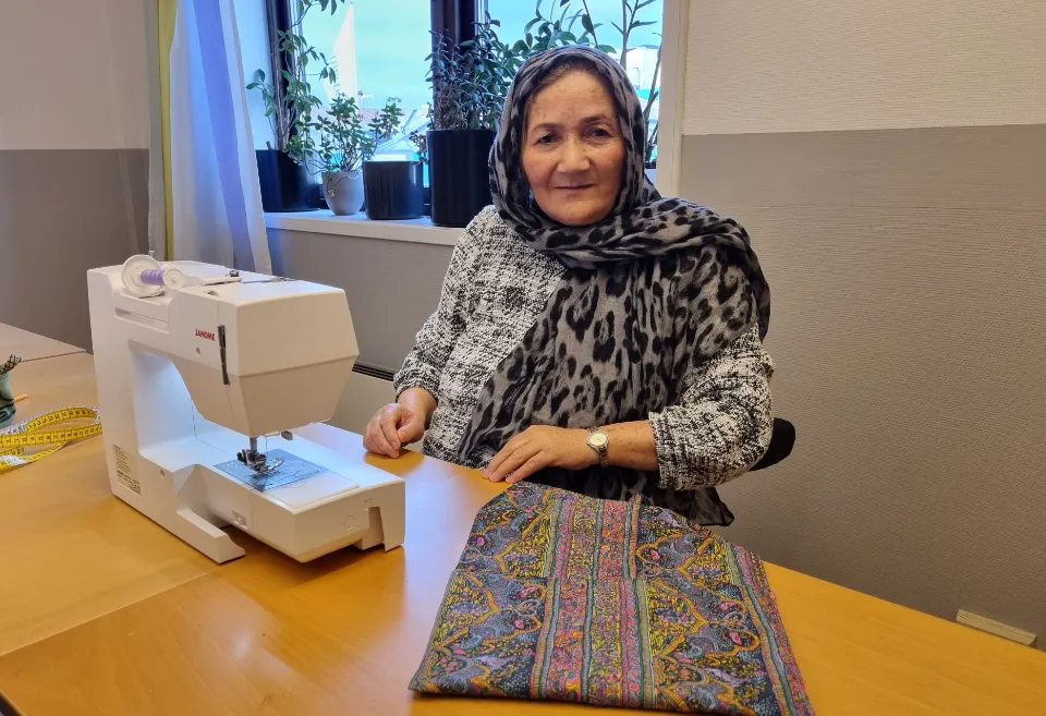 Kvinna med schal över håret, framför symaskin med ett tygstycke på bordet.