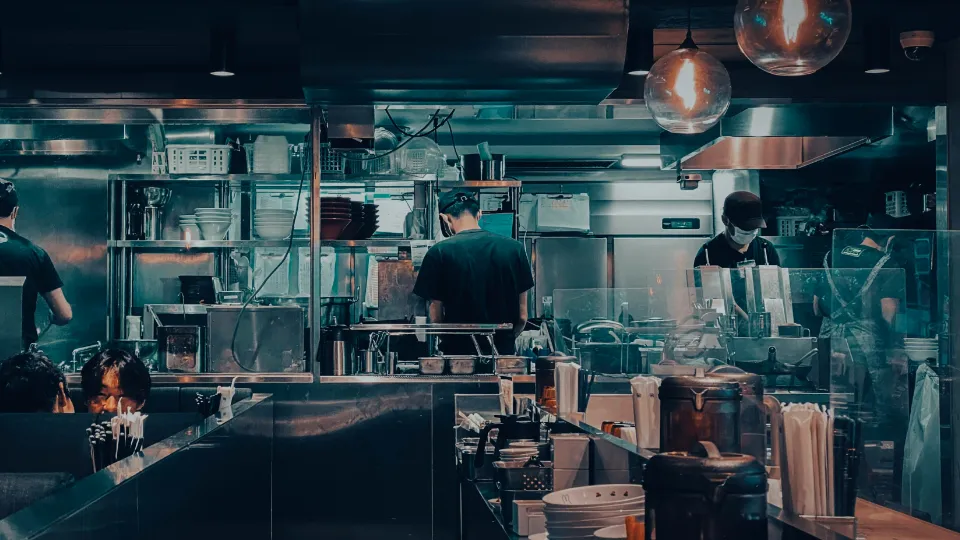 Samarbetet mellan arbetsliv och utbildning utreds i Finland Öppet restaurangkök med kockar som arbetar vid spisar och rostfria bänkar under hänglampor.