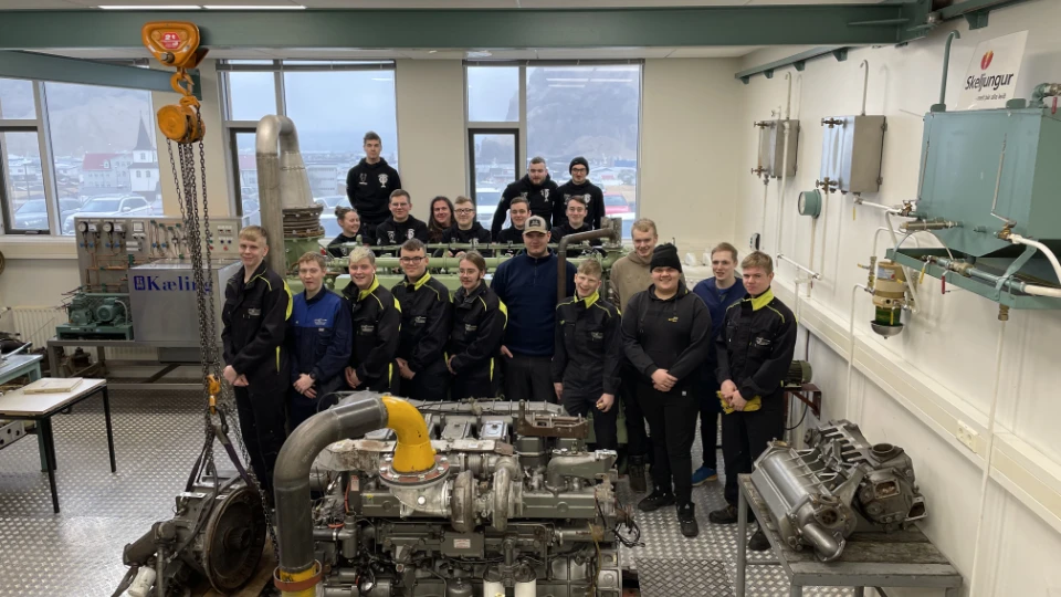 FÍV skolen studentene Gruppefoto av elever i verksted ved FÍV skole på Island, foran motor- og maskindeler og teknisk utstyr.