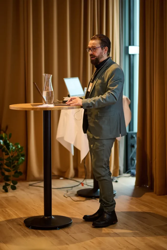 Brandur Hanusarson holder oplæg ved et ståbord med laptop og mikrofon under en konference i et indendørs lokale.