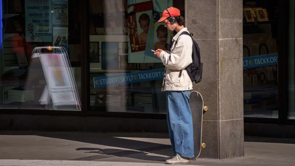 Elevteam inom komvux med fokus på unga vuxna i utanförskap Person med röd keps, hörlurar och ryggsäck står med skateboard utanför en butik och tittar på sin mobil.