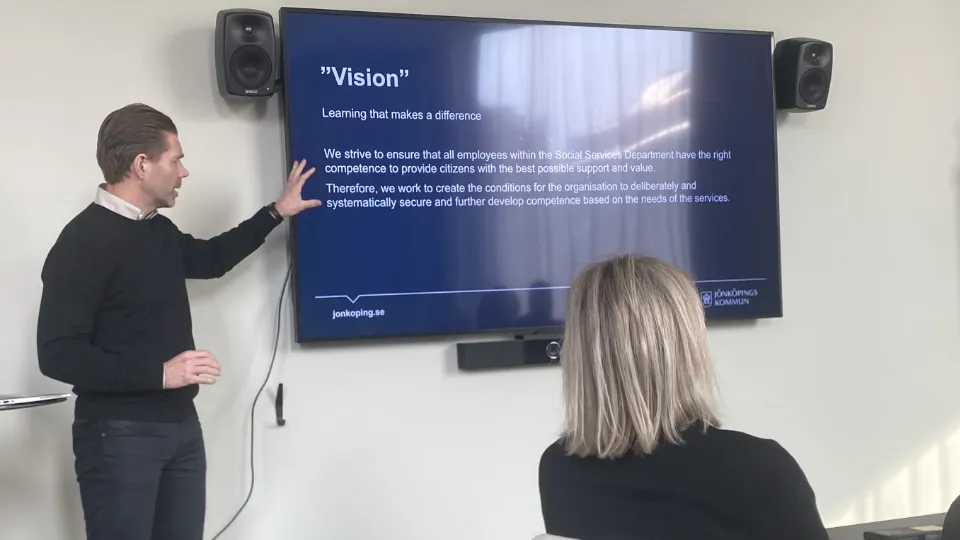 Martin Sinclair En mann holder en presentasjon foran en stor skjerm med teksten «Vision», mens en person sitter og ser på i et møterom.