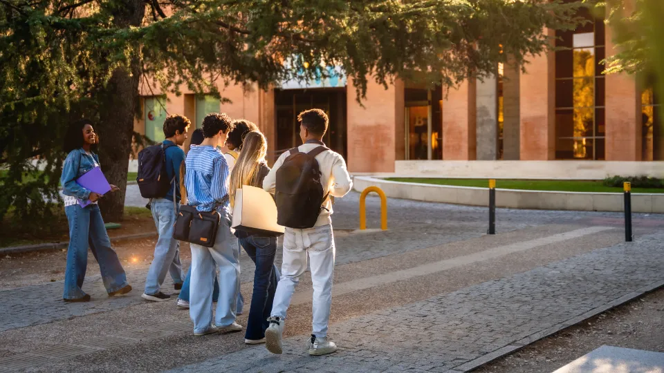 Snabbare väg till spetskompetens – Regeringen vill stärka yrkeshögskolan Studenter med ryggsäckar går på en campusväg framför en universitetsbyggnad i varmt eftermiddagsljus.