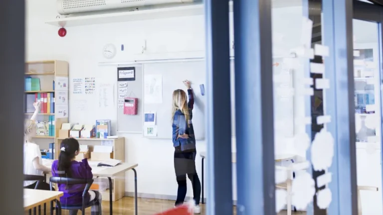 En lärare skriver på whiteboardtavlan framför elever i ett ljust klassrum.