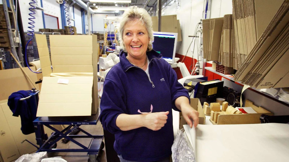 Kvinde på en pakkelinje i et produktionslokale omgivet af papkasser og emballagematerialer.