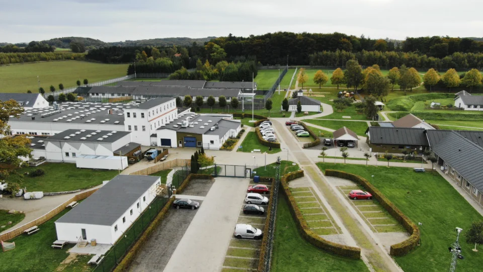 Luftfoto af Jyderup Fængsel med hvide bygninger, veje, parkeringspladser og grønne områder omgivet af skov. Fængslet er Danmarks eneste kvindefængsel.
