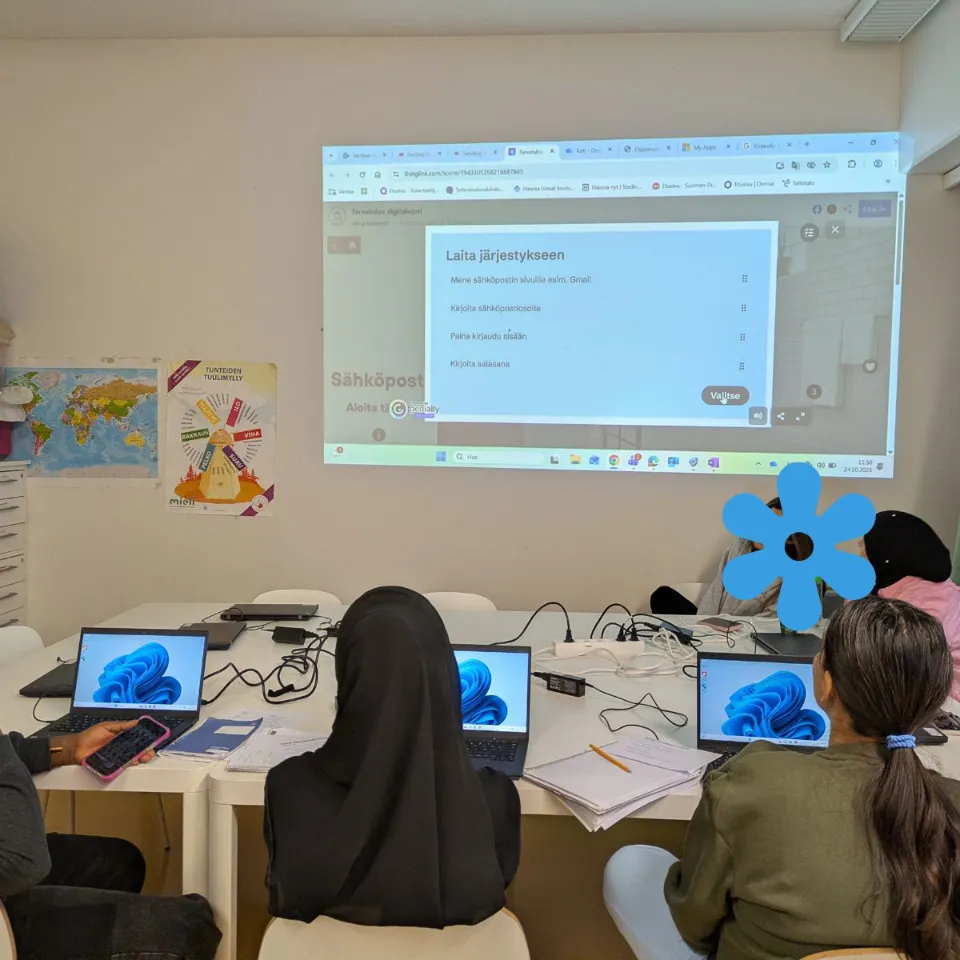 Personer sitter runt ett bord med bärbara datorer under en lektion, medan instruktioner om att skapa ett Gmail-konto visas på väggen med projektor.