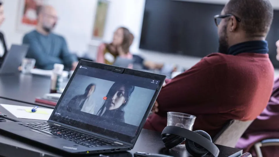 A laptop on a meeting table shows a video call, while people sit in a blurred conference room with headphones and a cup nearby.