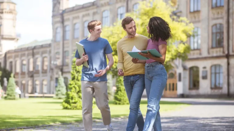 Tre studenter promenerar och samtalar på ett universitetsområde med mappar i händerna.
