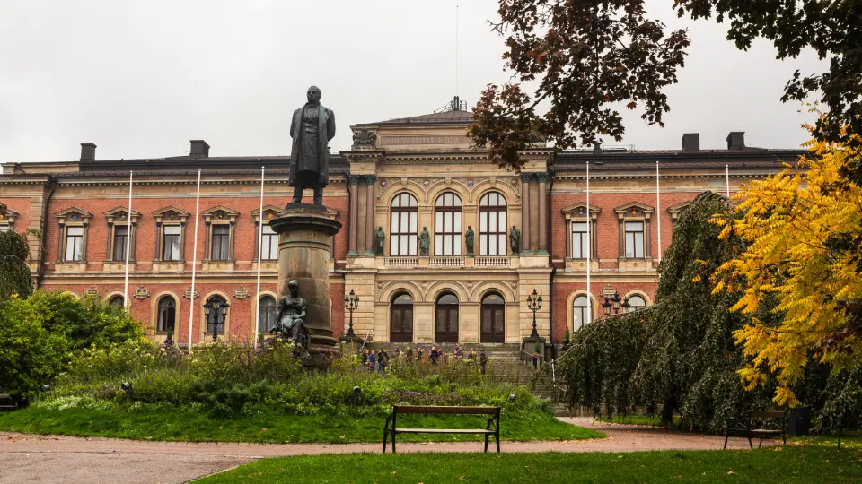 Historisk universitetsbyggnad med staty och grönskande park i förgrunden.