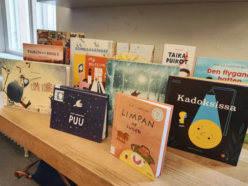 A collection of illustrated children's books, including wordless picture books, displayed on a wooden shelf at Turku City Library in Finland.
