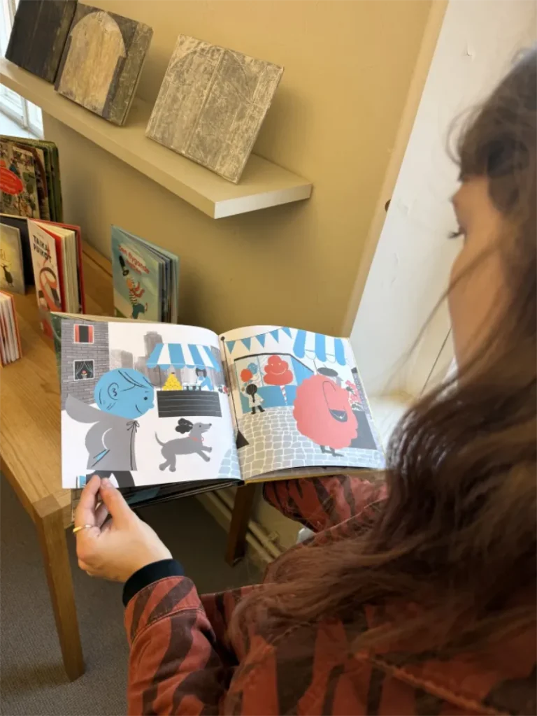 A woman explores a colorful picture book without words at a library, with children's books and artwork displayed on shelves behind her.