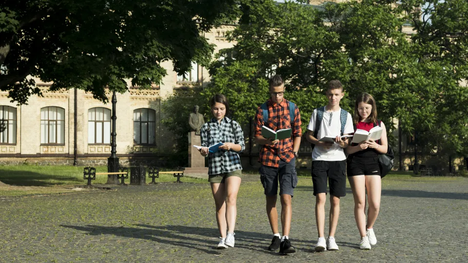 Four university students walking across campus while reading books, with a historic academic building and green trees in the background.