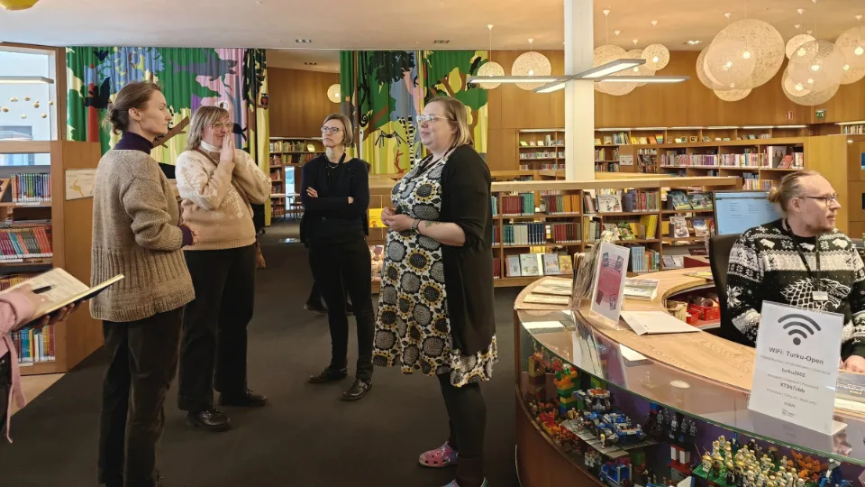 Participants in a study visit gather for a discussion inside Turku City Library, surrounded by bookshelves, colorful decorations, and a curved library service desk.