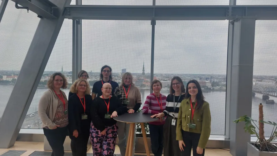 Eight participants from Finland, Lithuania, and Latvia pose for a group photo in front of large windows overlooking a city and river during a library study visit.