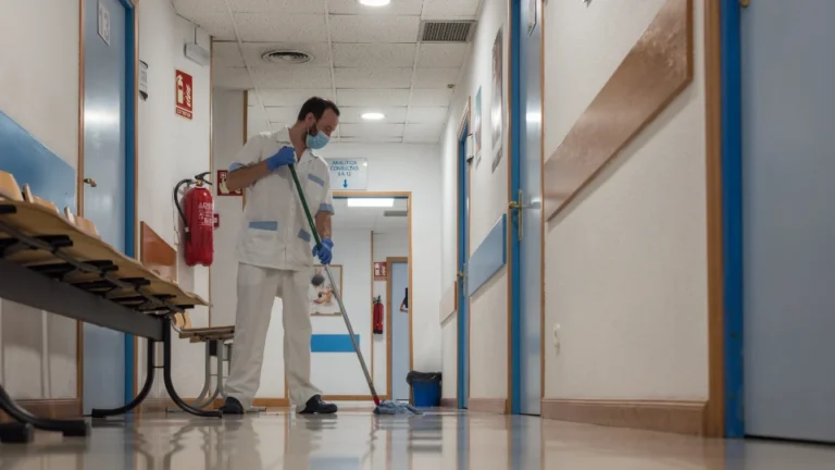 Rengøringsmedarbejder med mundbind og handsker vasker gulv på en hospitalsgang.